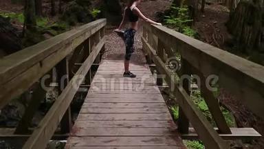 一个年轻女子穿着运动用品在一座常绿森林里的木桥上伸展着一条小溪的全景。
